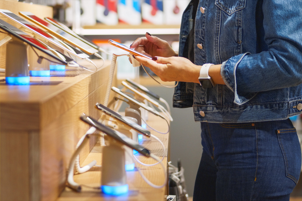 Person browsing smartphones on display at a store, with multiple devices secured on a wooden counter with illuminated stands.