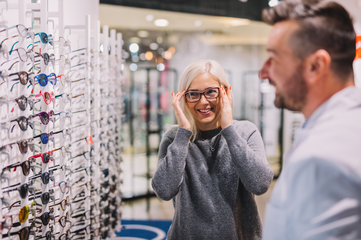 A woman tries on eyeglasses at an optical store while smiling and talking to a salesperson.