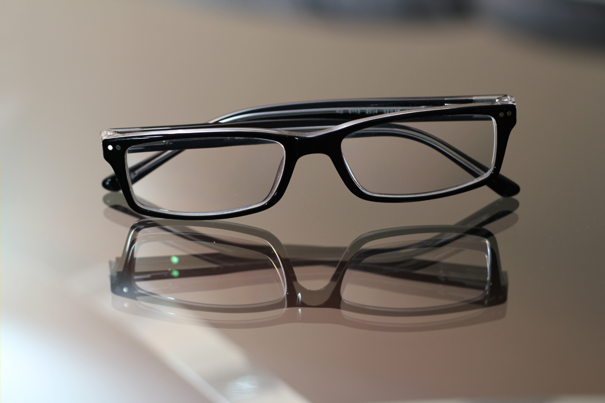 Black rectangular eyeglasses with clear temples placed on a reflective surface, showing their reflection.