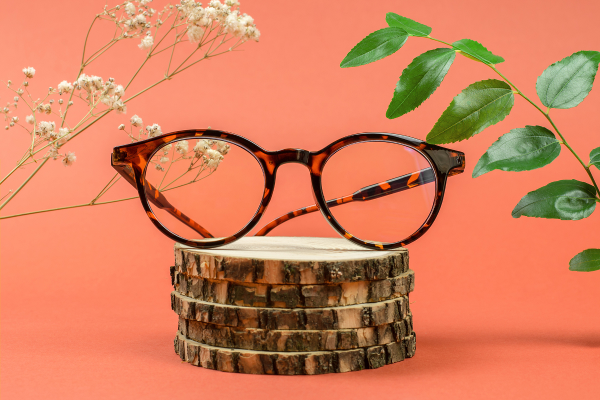 Tortoiseshell round eyeglasses displayed on stacked wooden pieces with flowers and green leaves in the background against a coral backdrop.