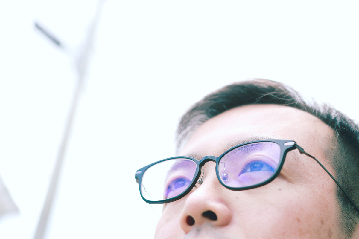 Close-up of a person wearing glasses and looking up with a bright sky in the background.