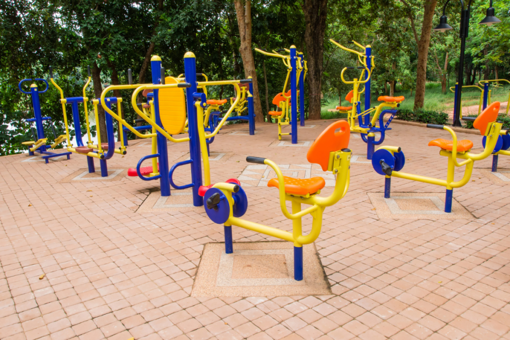 Colorful outdoor gym equipment with yellow and blue frames set on a tiled surface in a park surrounded by trees.