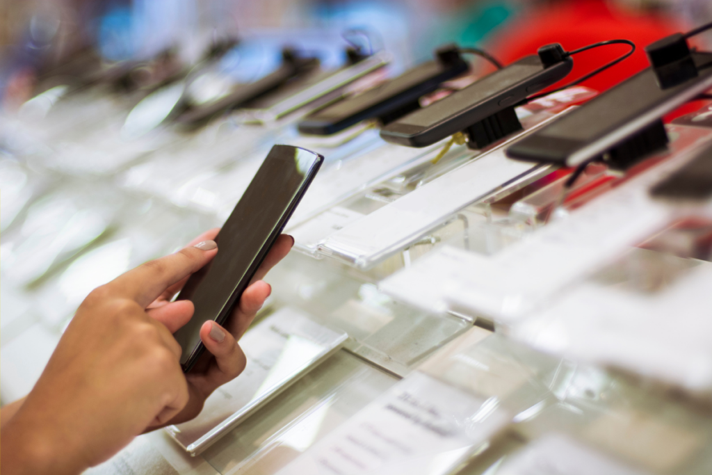 Person holding a smartphone while browsing a display of multiple devices in a retail store, with secured models on clear stands.