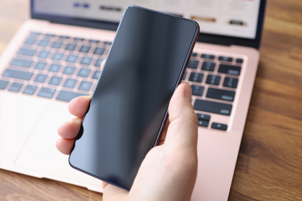 Person holding a smartphone with a blank screen in front of an open laptop on a wooden desk.