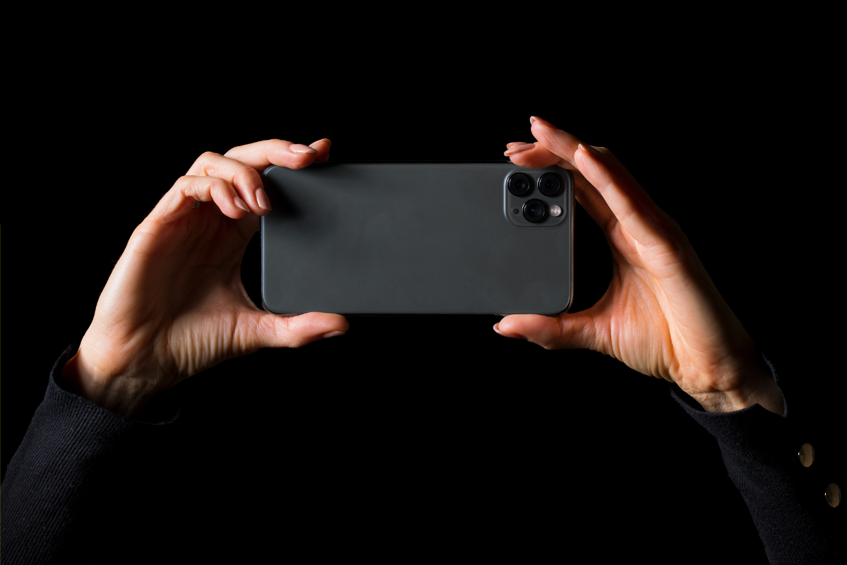 Person holding a smartphone horizontally against a black background, highlighting the rear camera setup.