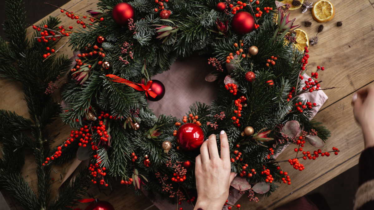 A beautifully crafted holiday wreath with lush green fir branches, bright red berries, golden accents, and ornaments. A hand is delicately arranging the decorations, adding a personal touch to the festive centerpiece.