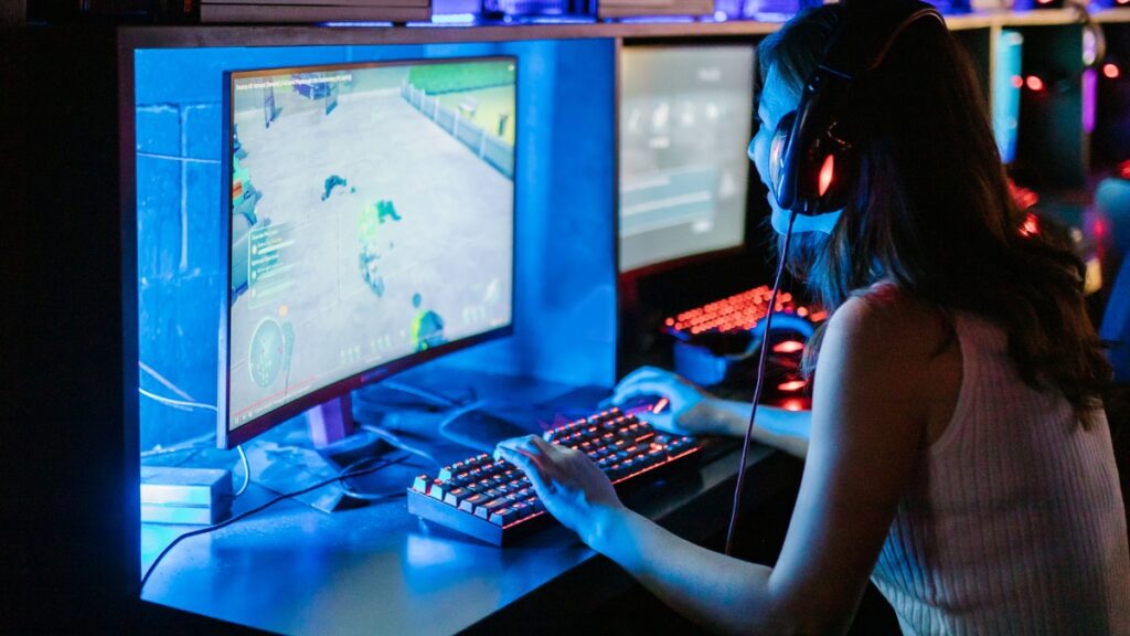 A gamer wearing headphones plays a PC game on a curved monitor with a glowing keyboard in a dimly lit gaming setup.
