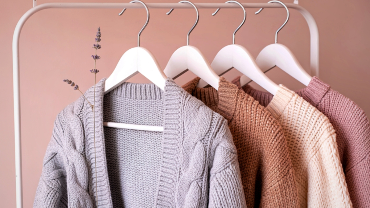 Neutral-toned knit sweaters hanging on white rack against pink wall